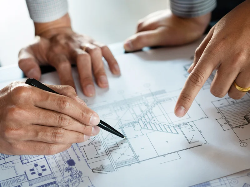 Hands on architectural blueprints; one writes with a pen while another points to a detail.