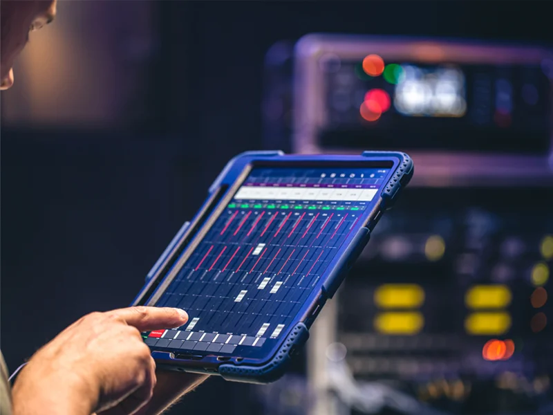 Close-up of a tablet showing a digital audio mixer interface, held by a hand, with blurred server racks in the background in a data-center setting.