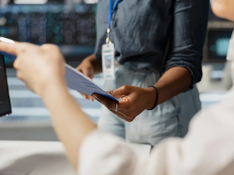 Person handing a blue folder to a coworker in a workspace, badge visible on a lanyard.
