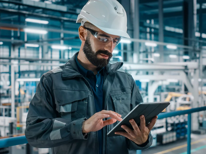 Industrial worker in a hard hat and safety goggles uses a tablet in a factory setting.